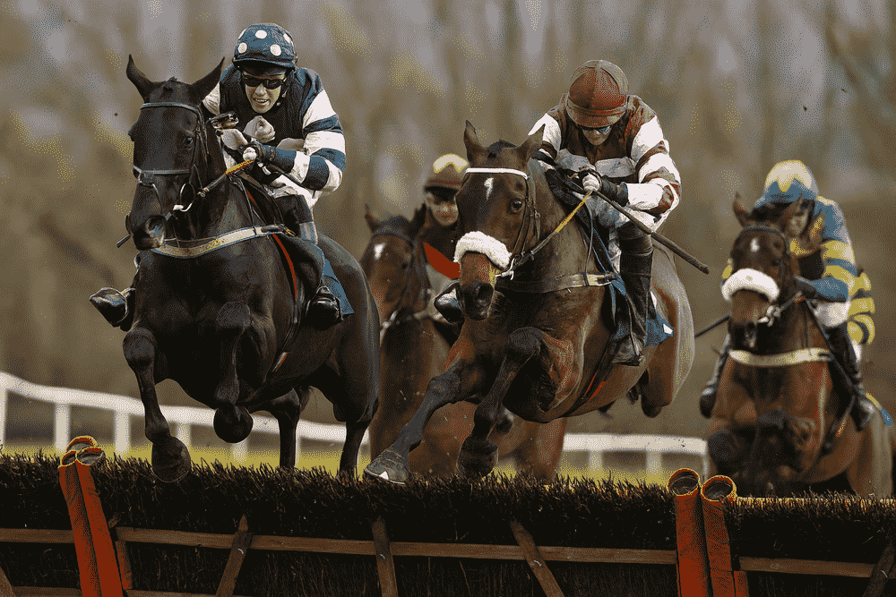 Several racehorses jumping a hurdle at Cheltenham during the 2025 Showcase, led by a jockey in green and orange silks mid-air during a National Hunt race