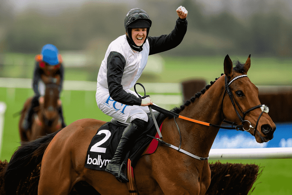 Jockey celebrating on a bay horse trained by Henry de Bromhead during a National Hunt jumps race, wearing black and white silks, representing the Knockeen yard.