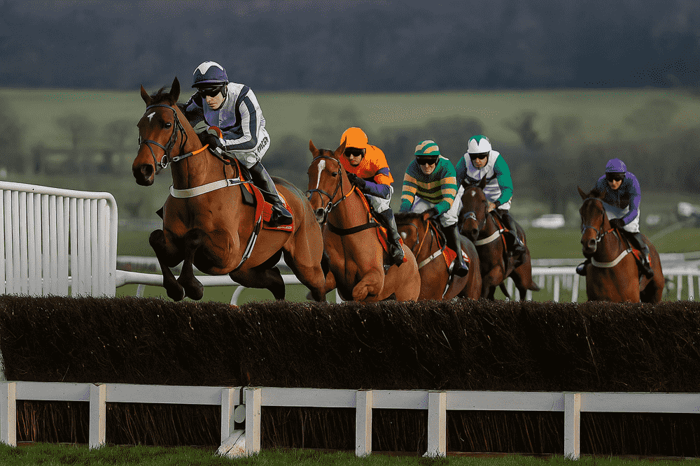 Five horses and jockeys jumping a steeplechase fence during a National Hunt race, with the leader in front and rivals chasing closely behind