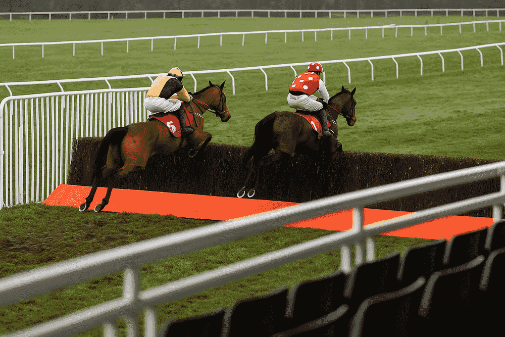 Two racehorses jumping a fence viewed from the grandstand during a winter National Hunt race, showing December jumping conditions
