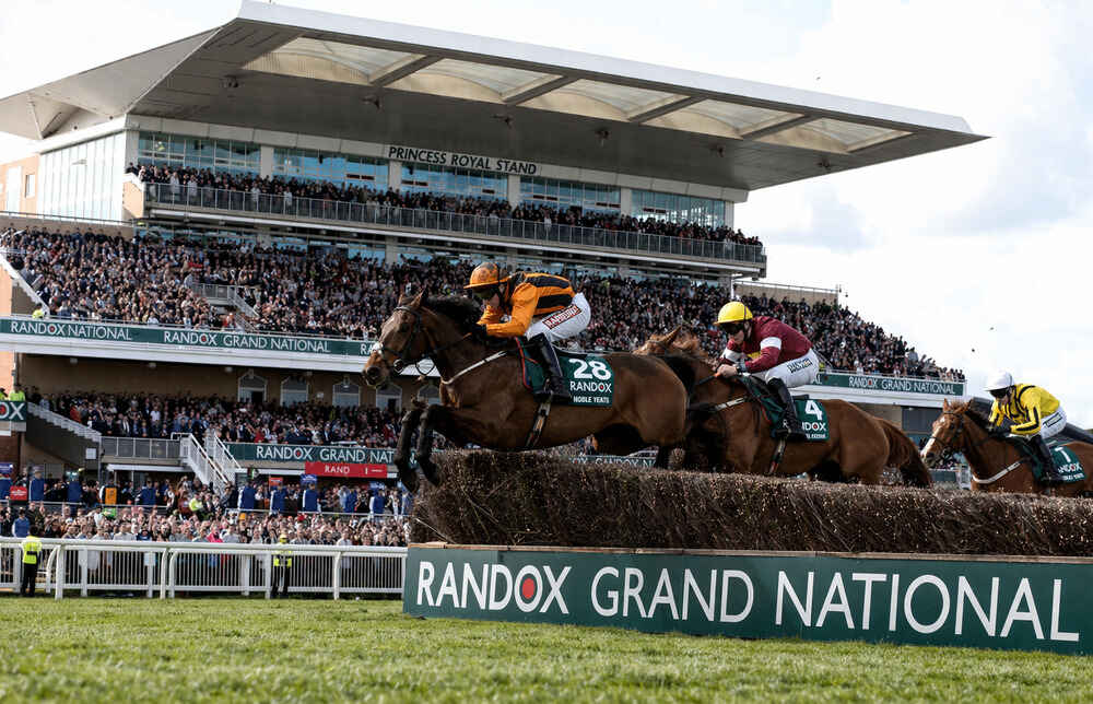 Grand National runners jump a fence at Aintree in front of the Princess Royal Stand and a packed crowd