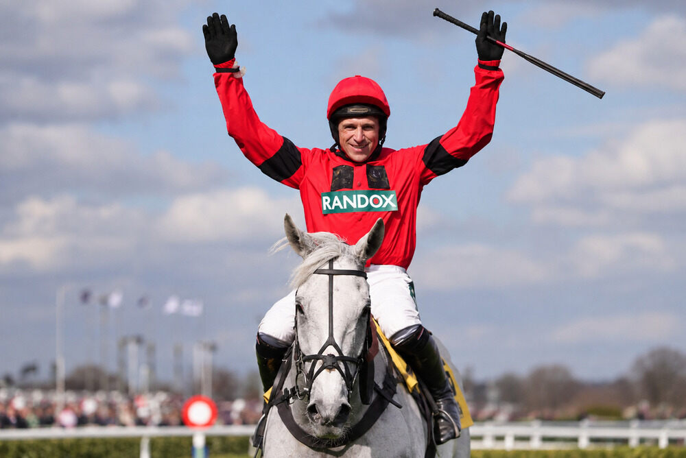 Harry Skelton raises both arms while riding a grey National Hunt racehorse.